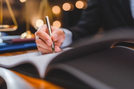 A lawyer in a suit writing at their desk, with the scales of justice behind them, representing legal work and consultation.