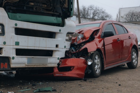 A red passenger car with severe front-end damage and debris on the ground after an accident involving a large white commercial truck.
